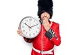 Middle age handsome wales guard man wearing traditional uniform holding big clock covering mouth with hand, shocked and afraid for