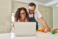 Middle age hispanic couple cooking and using laptop at kitchen Royalty Free Stock Photo