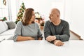 Middle age hispanic couple sitting on the table by christmas tree looking to side, relax profile pose with natural face with Royalty Free Stock Photo