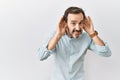 Middle age hispanic man with beard standing over isolated background trying to hear both hands on ear gesture, curious for gossip Royalty Free Stock Photo