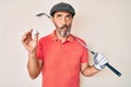 Middle age hispanic man holding golf club and ball making fish face with mouth and squinting eyes, crazy and comical