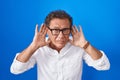 Middle age hispanic man standing over blue background trying to hear both hands on ear gesture, curious for gossip Royalty Free Stock Photo