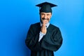 Middle age hispanic man wearing graduation cap and ceremony robe praying with hands together asking for forgiveness smiling