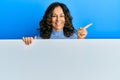 Middle age hispanic woman holding blank empty banner cheerful with a smile on face pointing with hand and finger up to the side