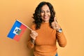 Middle age hispanic woman holding haiti flag smiling happy and positive, thumb up doing excellent and approval sign