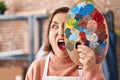 Middle age hispanic woman holding painter palette close to face angry and mad screaming frustrated and furious, shouting with