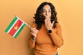 Middle age hispanic woman holding suriname flag covering mouth with hand, shocked and afraid for mistake