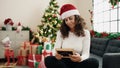 Middle age hispanic woman looking photo sitting on sofa by christmas tree at home