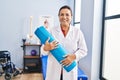 Middle age hispanic woman physiotherapist holding yoga mat at rehab clinic