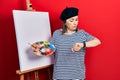 Middle age hispanic woman standing drawing with palette by painter easel stand checking the time on wrist watch, relaxed and