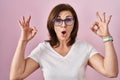 Middle age hispanic woman standing over pink background looking surprised and shocked doing ok approval symbol with fingers Royalty Free Stock Photo