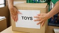 Middle age hispanic woman volunteer putting toys paper on cardboard box at charity center