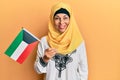 Middle age hispanic woman wearing hijab holding kuwait flag looking positive and happy standing and smiling with a confident smile