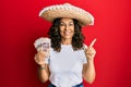 Middle age hispanic woman wearing mexican hat holding pesos banknotes smiling happy pointing with hand and finger to the side Royalty Free Stock Photo