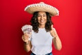 Middle age hispanic woman wearing mexican hat holding pesos banknotes smiling happy and positive, thumb up doing excellent and Royalty Free Stock Photo