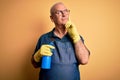 Middle age hoary cleaner man cleaning wearing apron and gloves using sprayer serious face thinking about question, very confused