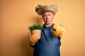 Middle age hoary farmer man wearing apron and hat holding plant pot over yellow background pointing with finger to the camera and