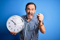 Middle age hoary man holding big clock standing over isolated blue background annoyed and frustrated shouting with anger, crazy Royalty Free Stock Photo
