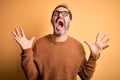Middle age hoary man wearing brown sweater and glasses over isolated yellow background crazy and mad shouting and yelling with