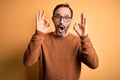 Middle age hoary man wearing brown sweater and glasses over isolated yellow background looking surprised and shocked doing ok