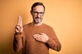 Middle age hoary man wearing brown sweater and glasses over isolated yellow background smiling swearing with hand on chest and