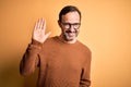 Middle age hoary man wearing brown sweater and glasses over isolated yellow background Waiving saying hello happy and smiling,
