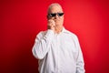 Middle age hoary man wearing funny sunglasses over isolated red background with hand on chin thinking about question, pensive