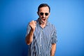 Middle age hoary man wearing striped shirt and sunglasses over isolated blue background angry and mad raising fist frustrated and Royalty Free Stock Photo