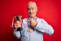 Middle age hoary successful man wearing medals holding trophy over isolated red background very happy pointing with hand and