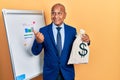 Middle age latin man wearing business suit holding dollars bag pointing thumb up to the side smiling happy with open mouth Royalty Free Stock Photo