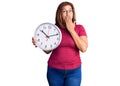 Middle age latin woman holding big clock covering mouth with hand, shocked and afraid for mistake