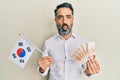 Middle age man with beard and grey hair holding south korea flag and won banknotes making fish face with mouth and squinting eyes, Royalty Free Stock Photo