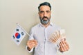 Middle age man with beard and grey hair holding south korea flag and won banknotes relaxed with serious expression on face Royalty Free Stock Photo