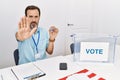 Middle age man with beard sitting by ballot holding i vote badge doing stop sing with palm of the hand