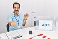 Middle age man with beard sitting by ballot holding i vote badge smiling cheerful offering palm hand giving assistance and