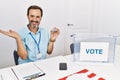 Middle age man with beard sitting by ballot holding i vote badge smiling cheerful presenting and pointing with palm of hand