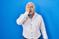 Middle age man with grey hair standing over blue background yawning tired covering half face, eye and mouth with hand Royalty Free Stock Photo