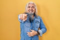 Middle age man with grey hair standing over yellow background laughing at you, pointing finger to the camera with hand over body, Royalty Free Stock Photo