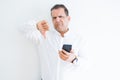 Middle age man using smartphone over white wall with angry face, negative sign showing dislike with thumbs down, rejection concept Royalty Free Stock Photo