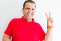 Middle age man wearing red t-shirt over white wall smiling with happy face winking at the camera doing victory sign Royalty Free Stock Photo