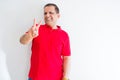 Middle age man wearing red t-shirt over white wall smiling looking to the camera showing fingers doing victory sign Royalty Free Stock Photo