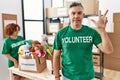 Middle age man wearing volunteer t shirt at donations stand showing and pointing up with fingers number three while smiling