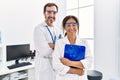 Middle age man and woman partners wearing scientist uniform holding clipboard with arms crossed gesture at laboratory