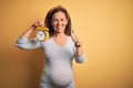 Middle age pregnant woman expecting baby at aged pregnancy holding alarm clock happy with big smile doing ok sign, thumb up with Royalty Free Stock Photo