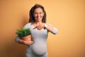 Middle age pregnant woman expecting baby holding plant pot very happy pointing with hand and finger Royalty Free Stock Photo