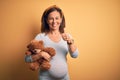 Middle age pregnant woman expecting baby holding teddy bear stuffed animal happy with big smile doing ok sign, thumb up with Royalty Free Stock Photo