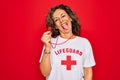 Middle age senior summer lifeguard woman holding whistle over red background sticking tongue out happy with funny expression