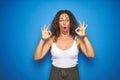 Middle age senior woman with curly hair standing over blue isolated background looking surprised and shocked doing ok approval Royalty Free Stock Photo