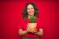 Middle age senior woman holding green cactus over red isolated background with a happy face standing and smiling with a confident Royalty Free Stock Photo