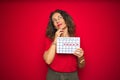 Middle age senior woman holding menstruation calendar over red isolated background serious face thinking about question, very